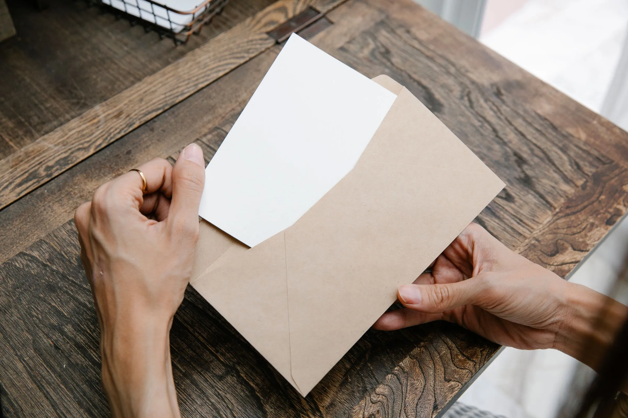 Hands sliding a letter out of a brown envelope on a wooden table.