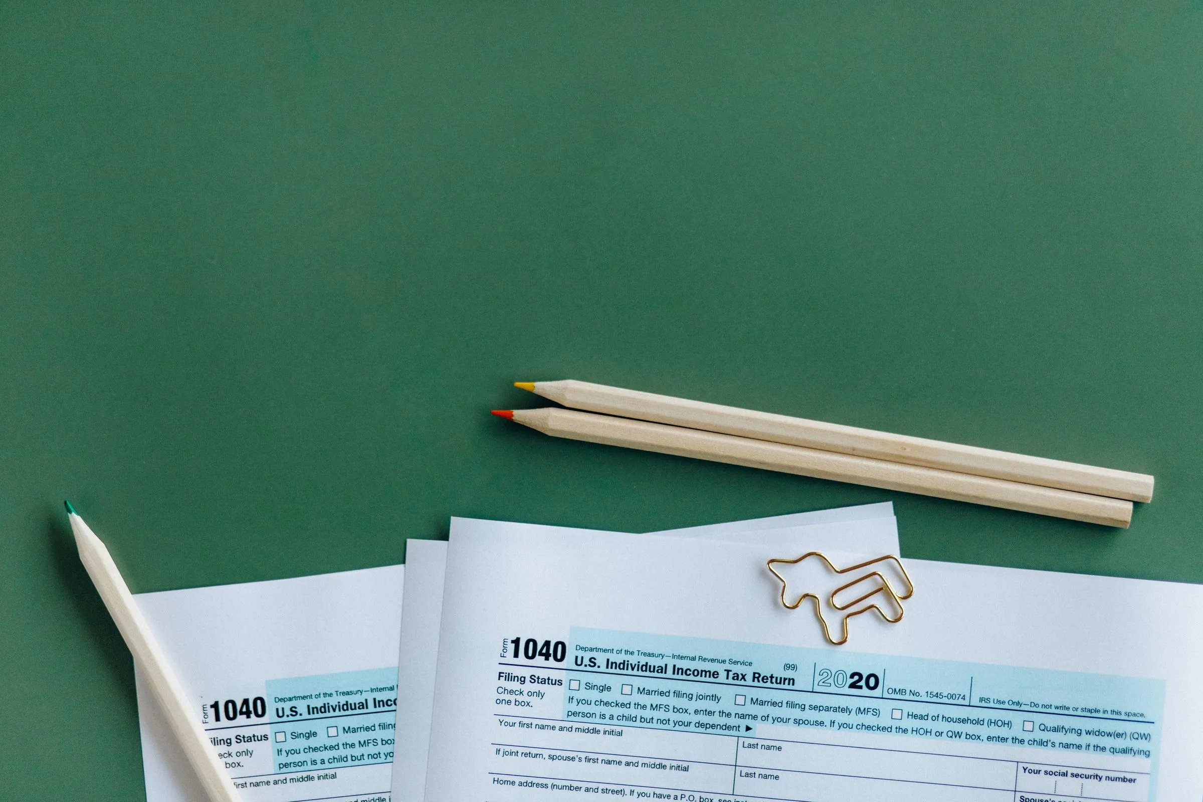 Blank Form 1040 tax returns on a green desk beside two pencils and paper clips.